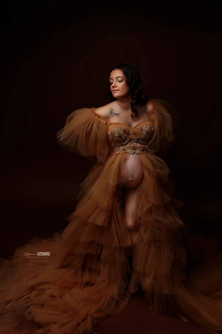 Femme enceinte posant en studio dans une robe volumineuse en tulle caramel ornée de broderies florales, photographie artistique par Stéphanie Azemard.