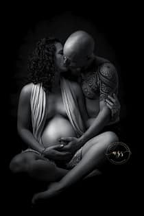 Photographie en noir et blanc d'une femme enceinte assise en tailleur avec son mari l'embrassant en studio à Vergèze.