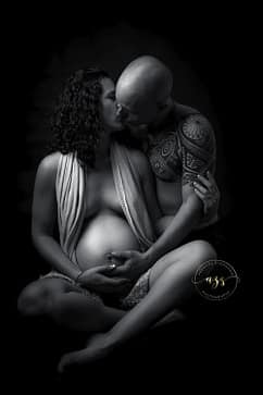 Photographie en noir et blanc d'une femme enceinte assise en tailleur avec son mari l'embrassant en studio à Vergèze.