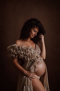 Femme enceinte vêtue d’une robe dorée scintillante, posant avec douceur et élégance devant un fond marron chaleureux. Une séance maternité capturant l’émotion et la beauté de la grossesse.