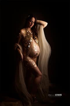 Femme enceinte posant avec grâce dans un studio, parée de feuilles d’or et d’un voile fluide, créant une ambiance élégante et artistique.