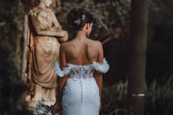 Vue de dos d'une mariée dans une robe élégante ornée de dentelle, tenant un bouquet, devant une statue classique, capturant un moment intemporel de grâce et de beauté.