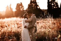 Un couple de mariés s’embrasse tendrement dans un champ baigné par la lumière dorée du coucher du soleil. La mariée porte une robe blanche fluide, tandis que le marié est vêtu d’un costume beige. L’environnement naturel, avec ses herbes hautes et ses fleurs rouges, ajoute une ambiance romantique et bucolique.