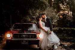 Un couple de jeunes mariés s'enlace tendrement devant une Ford Mustang dans une forêt romantique, baignée de lumière dorée.