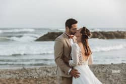 Un couple de mariés s’embrasse tendrement sur une plage, avec l’océan et des rochers en arrière-plan. La mariée porte une robe blanche à manches longues et un dos nu orné de fleurs, tandis que le marié est vêtu d’un costume beige et d’un nœud papillon.