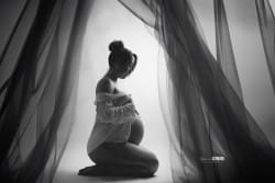 Portrait en noir et blanc d'une femme enceinte, agenouillée et entourée de voilages. Elle pose avec douceur, en tenant son ventre, symbole de maternité et de tendresse.