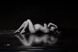 Femme enceinte allongée sur le côté, nue, dans une pose artistique et apaisée, capturée en noir et blanc avec un reflet dans l'eau, dans un style photographique élégant et minimaliste.