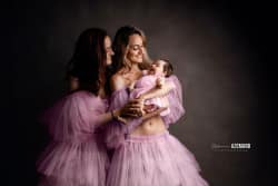 Deux femmes en robes en tulle rose tiennent avec tendresse un bébé habillé en rose, partageant un moment de complicité et d’amour lors d’une séance photo artistique sur fond sombre.