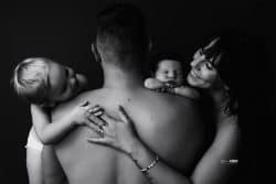 Une photo en noir et blanc d'une famille. Un homme de dos porte deux enfants sur ses épaules tandis qu'une femme sourit tendrement en le tenant par l'épaule.