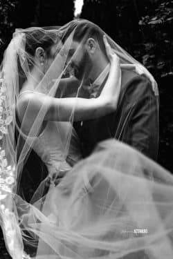 Un couple de mariés s'enlace tendrement sous un voile délicat, capturé en noir et blanc pour un effet intemporel.