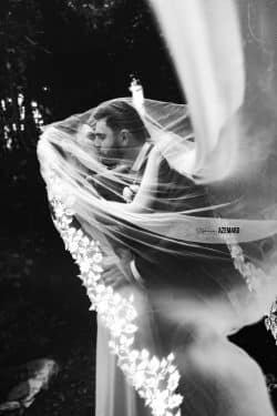 Un couple de mariés s’embrasse passionnément, enveloppé dans un voile de dentelle flottant, photographié en noir et blanc par Stéphanie Azémard.