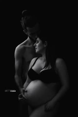 Portrait en noir et blanc d'un couple en attente d'un enfant, mettant en avant la tendresse et l'amour entre les futurs parents, avec un éclairage doux et un fond sombre.