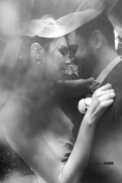 Portrait en noir et blanc d’un couple de mariés tête contre tête, enveloppés dans le voile de la mariée, photographié par Stéphanie Azémard.