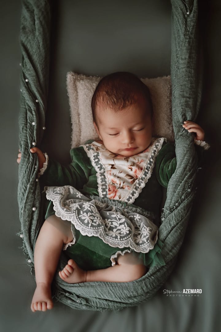 Un bébé endormi dans une balancelle suspendue, habillé d’une robe verte ornée de dentelle blanche et de fleurs, dans un décor studio élégant avec perles décoratives.
