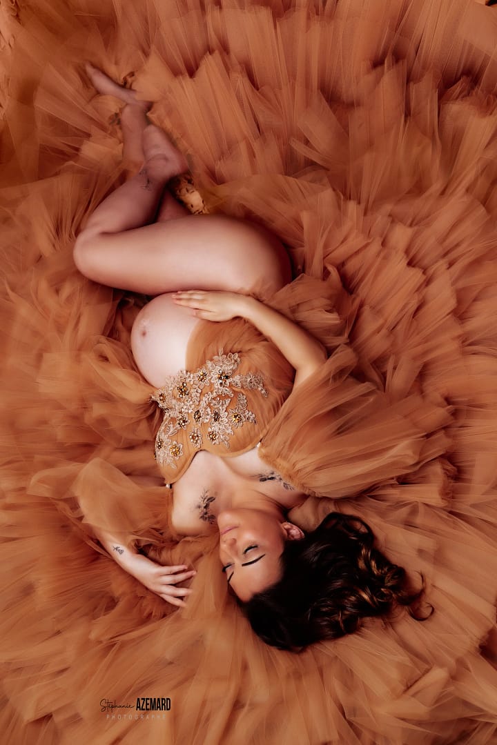 Femme enceinte allongée en robe de tulle caramel dans un studio photo, regard paisible, photographie artistique par Stéphanie Azemard.